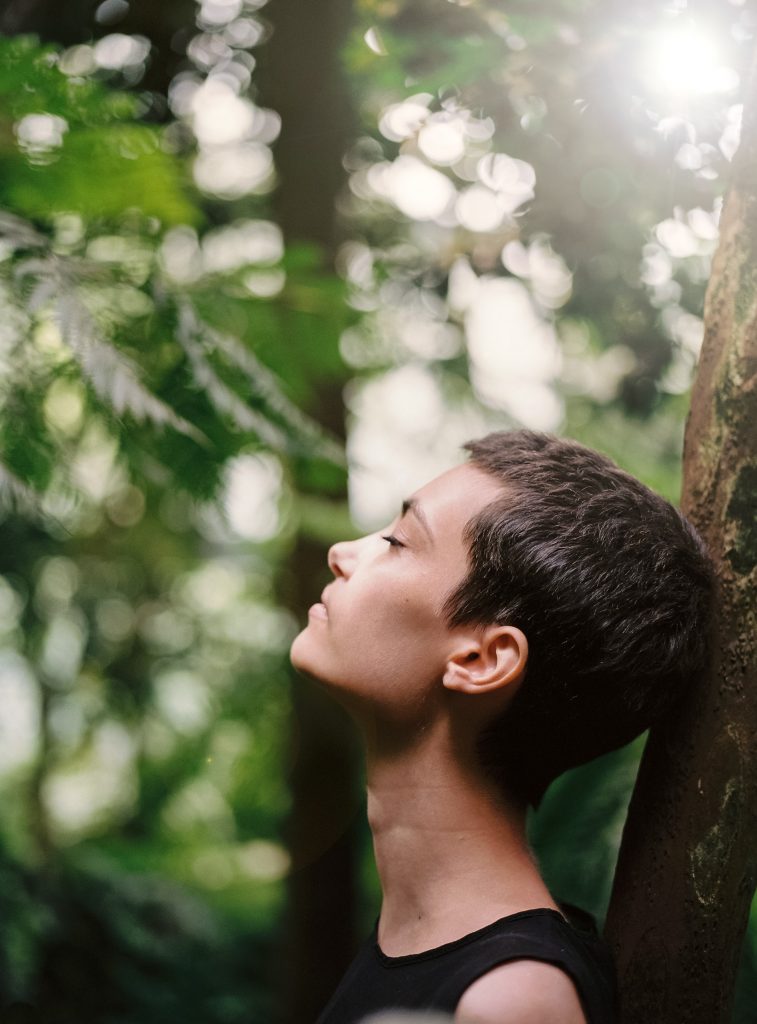 Young woman with short hair, eyes closed, head calmly leaning back against the trunk of a tree in the forest.