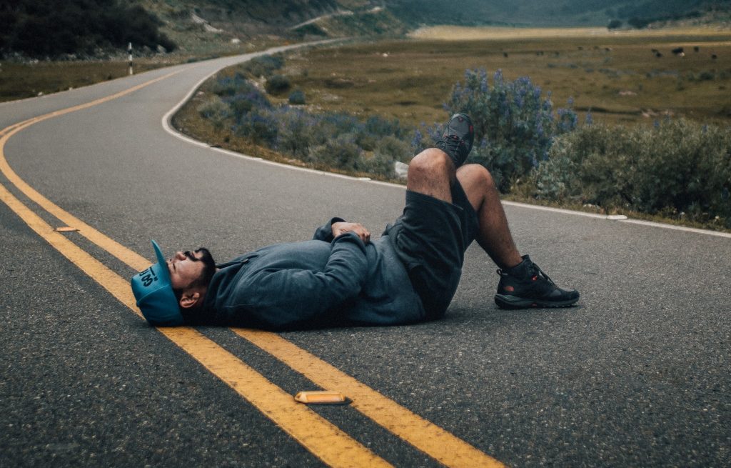 Bearded man in shorts and running shoes lying on his back with his feet standing and legs crossed in the middle of a paved road in an isolated valley surrounded by mountains