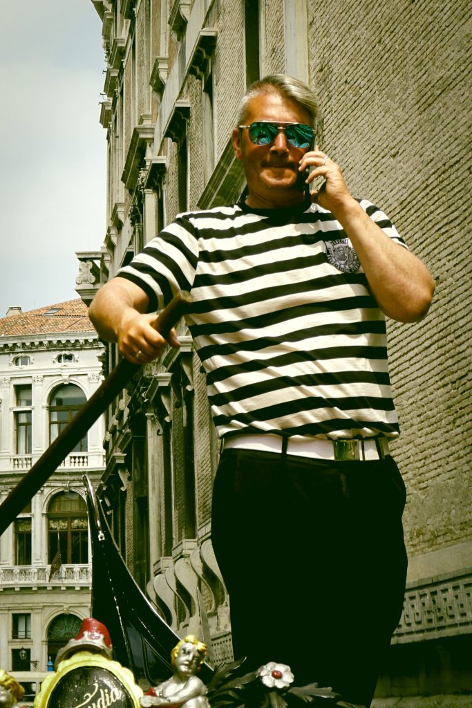 Blonde gondolier in Venice with mirrored shades wearing a black and white horizontally striped short sleeved shirt and a white belt on his black pants guiding his gondola while talking on his cell phone.