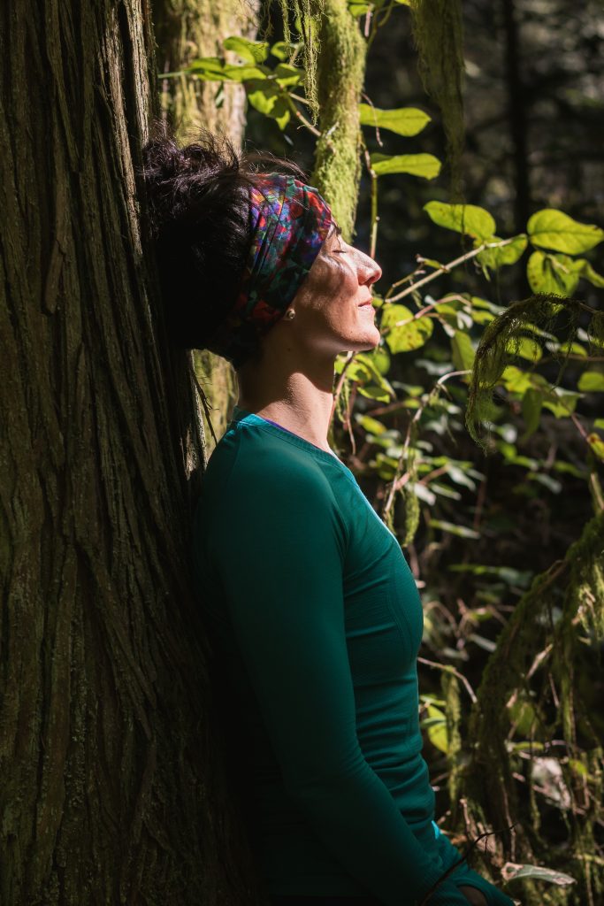 Middle aged woman with eyes closed resting against large tree in the forest. Do we notice our movements more with our eyes open or closed? Or with our minds and hearts open or closed?