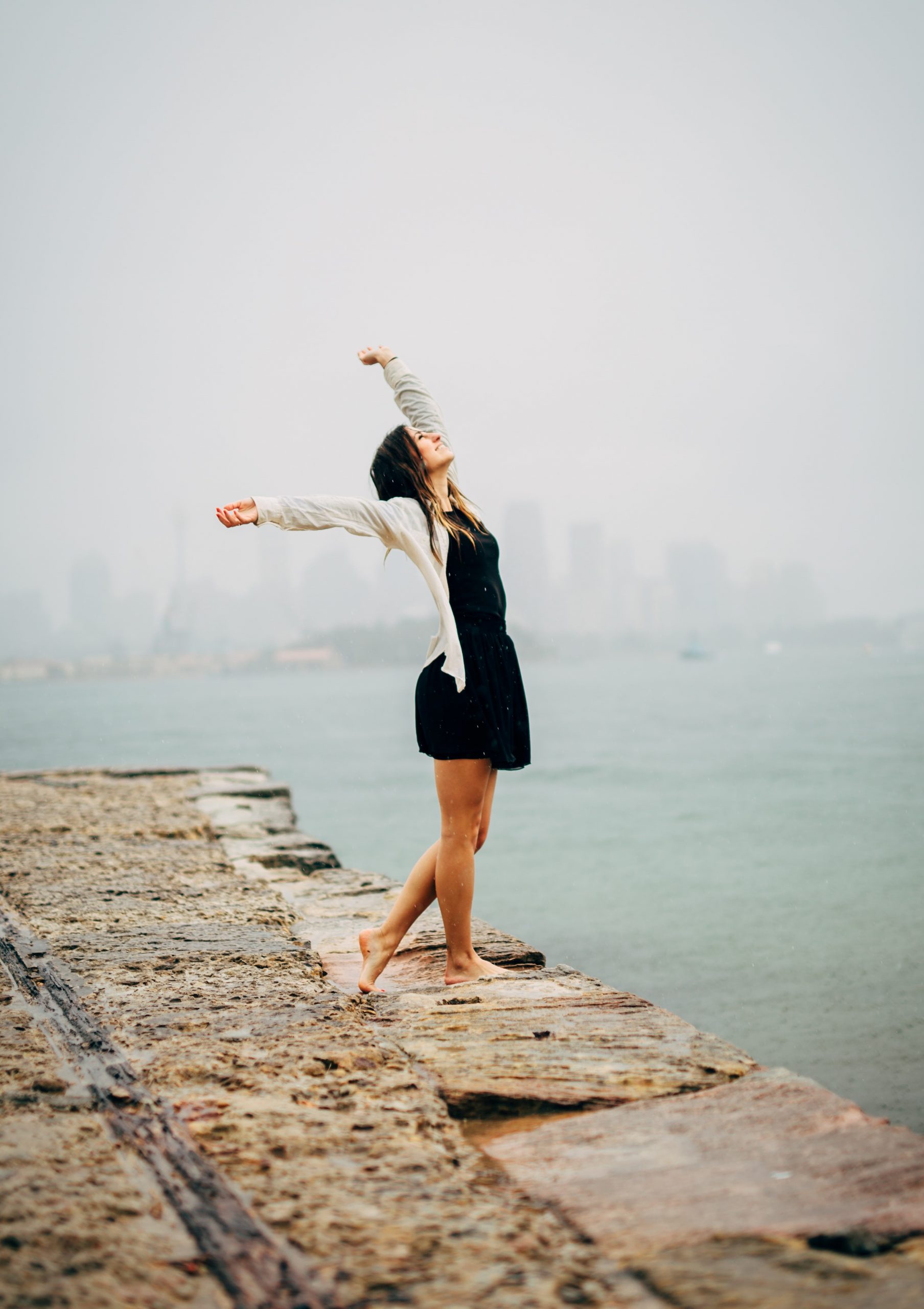 Young woman on a wall, outline of a city across the water in the background. She is reaching up and out, leaning back, looking up, opening herself to the world. Freeing Your Body brings this kind of joy.