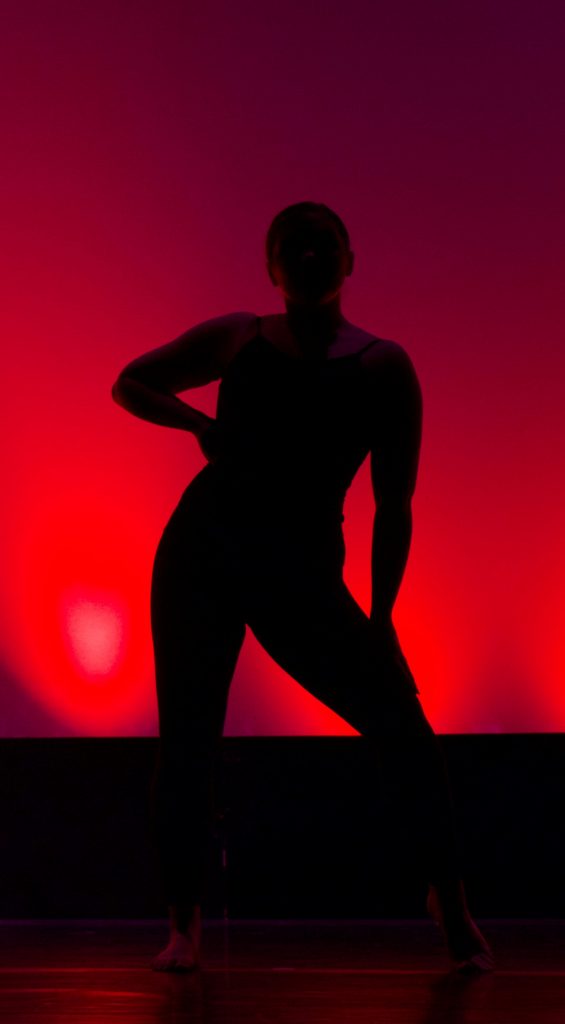 Silhouette of woman against red sunset-like backdrop with one hand on her extended hip as she leans the other way in an attitude of confidence. Freeing Your Hips makes it easy and natural to express yourself with your whole body.