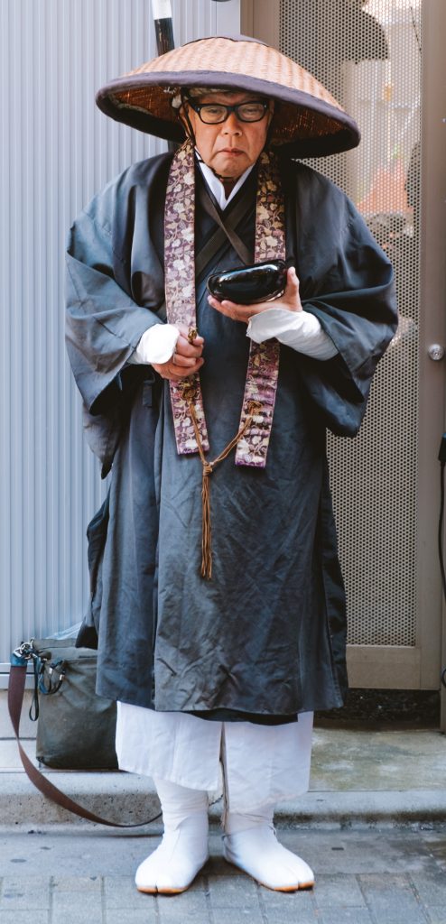 Elderly Asian priest in traditional dress: white footwear, loose white pants, long grey robe, multi-colored scarf hanging down his chest, wide brimmed straw hat, holding a small bowl.
