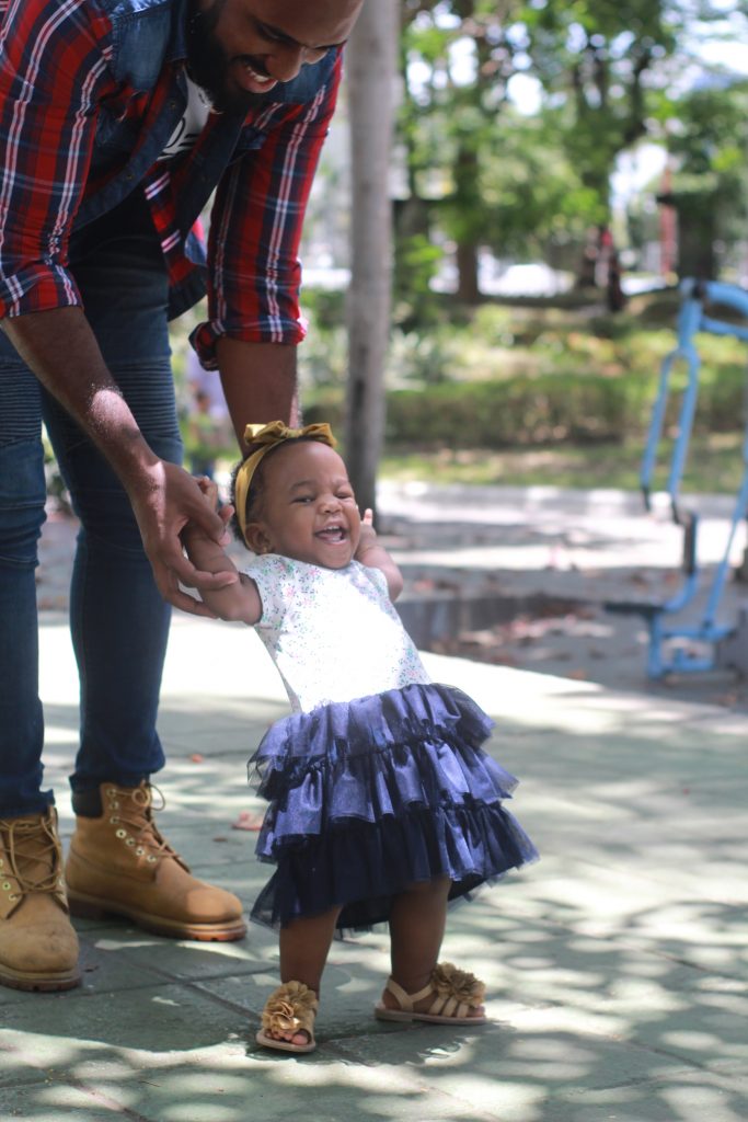 Excited toddler taking tentative steps as her father holds her hands. Baby steps take us a long way.