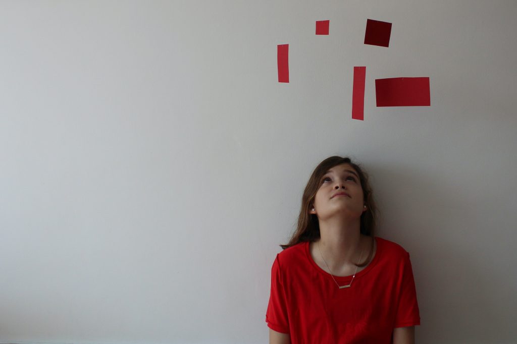 Young girl in red against white wall looking up at a mobile of red rectangles of various sizes and dimensions. Mindfulness is noticing with your imagination.