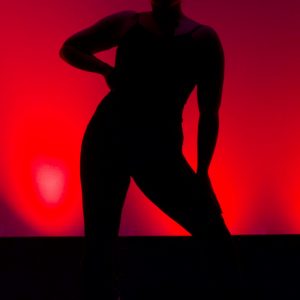 Silhouette of woman against red sunset-like backdrop with one hand on her extended hip as she leans the other way in an attitude of confidence. Freeing Your Hips makes it easy and natural to express yourself with your whole body.