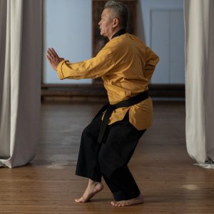 Man in ochre top with black sash and black pants doing Tai Chi. He is lifting his right foot gracefully, well grounded on his left foot, bending both his knees, his pelvis stable and centered.