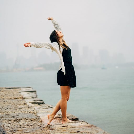 Young woman on a wall, outline of a city across the water in the background. She is reaching up and out, leaning back, looking up, opening herself to the world. Freeing Your Body brings this kind of joy.