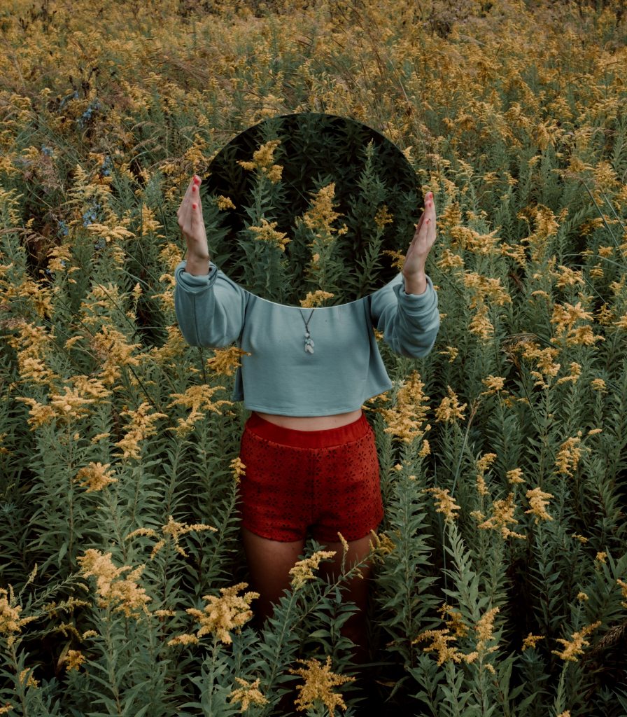 Woman standing in a field of flowers holding a circular image of the flowers in front of her face, which blends into the flowers in the field so she has no face to identify her.
