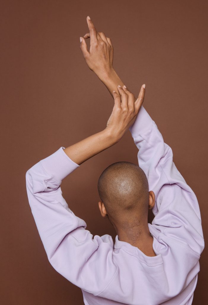 Person with very short black hair, in loose, comfortable lavender shirt, back turned, first fingers extended, right arm raised easily above head and left arm gracefully bent to encircle head.