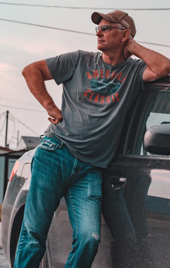 Older man in well worn jeans and t-shirt leaning comfortably against his car, his pelvis tilting easily one way while his shoulders tilt the other, one hand on hip, his head leaning against the other.