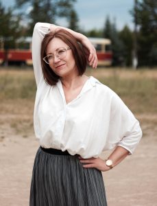 Smiling middle aged woman in a white top and grey skirt with her right arm draped gracefully over her head and her left hand on her tilted hip.