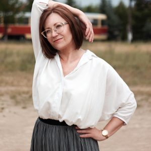 Smiling middle aged woman in a white top and grey skirt with her right arm draped gracefully over her head and her left hand on her tilted hip.